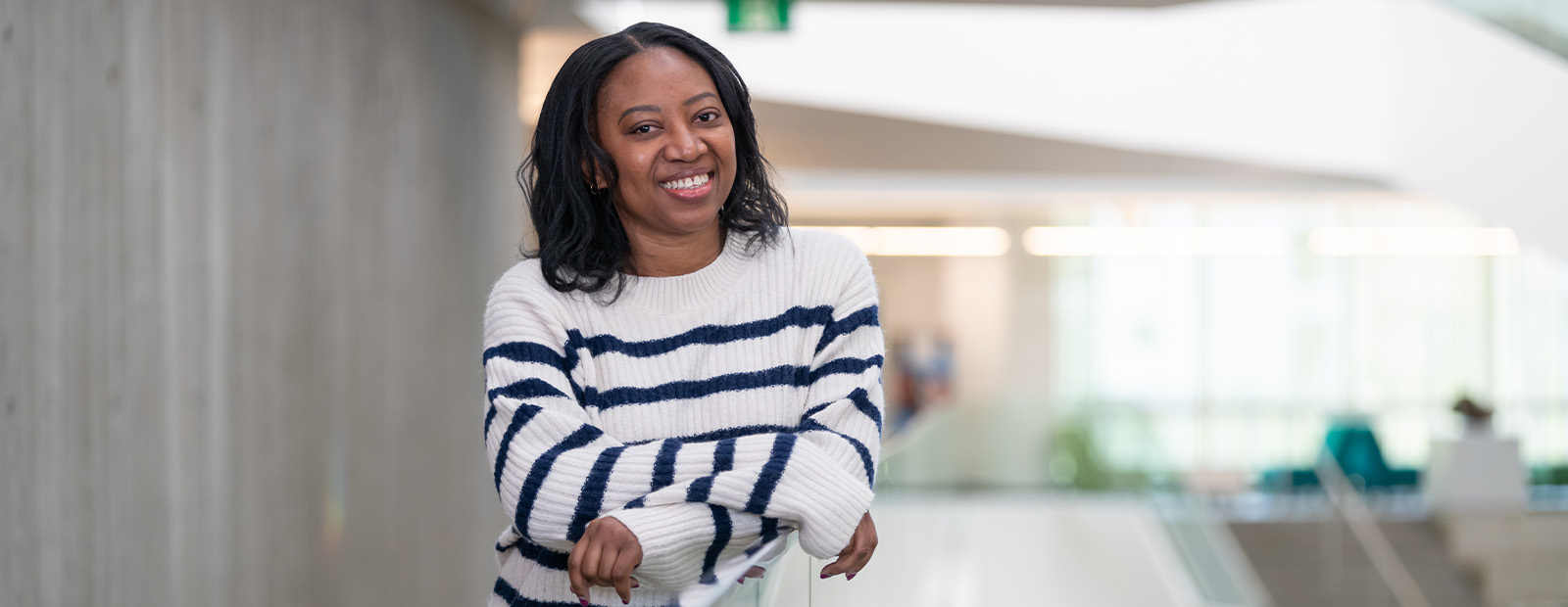 Tracy Thomas wears a white sweater with thin black horizontal stripes. She leans against a glass railing in Allard Hall, smiling.