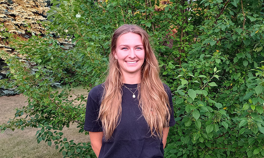 Shannon Majeau stands, smiling, in front of greenery