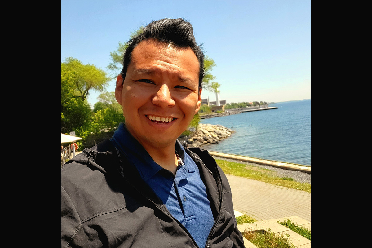 A man smiles at the camera while standing in front of a lake