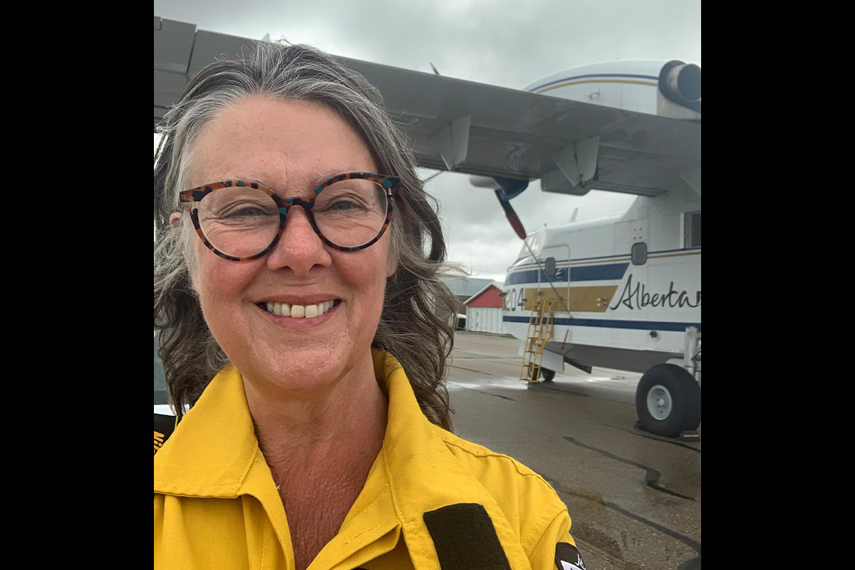 A woman with grey hair and tortoiseshell glasses takes a selfie in front of a small aircraft with the Government of Alberta wordmark on it