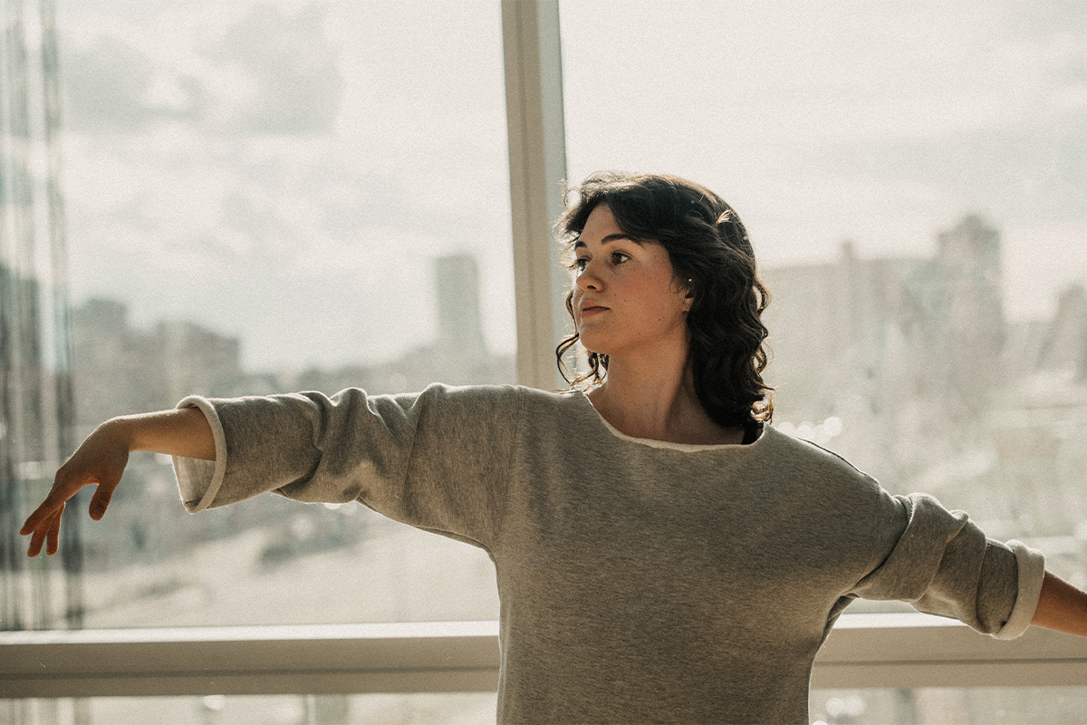 A woman with curly brown hair dances in front of a window