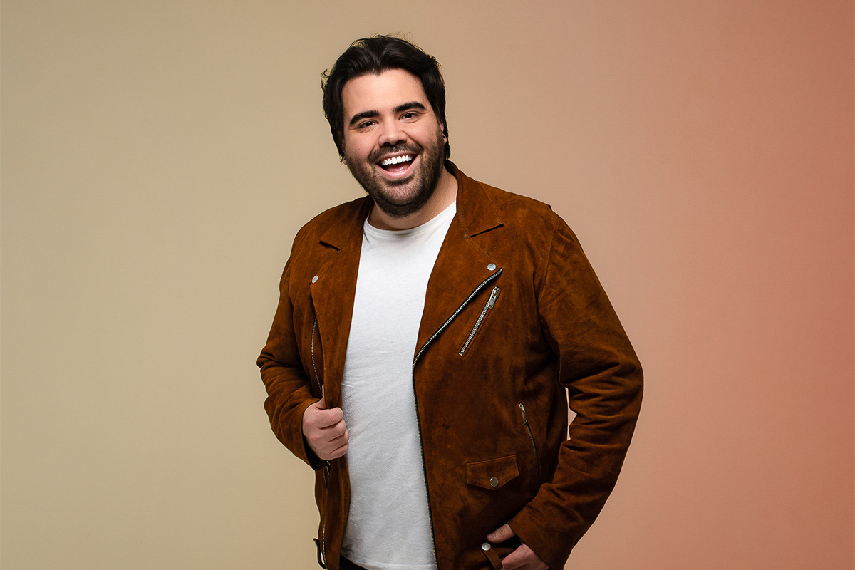 A man in a red velvet jacket smiles at the camera in front of a brown gradient background