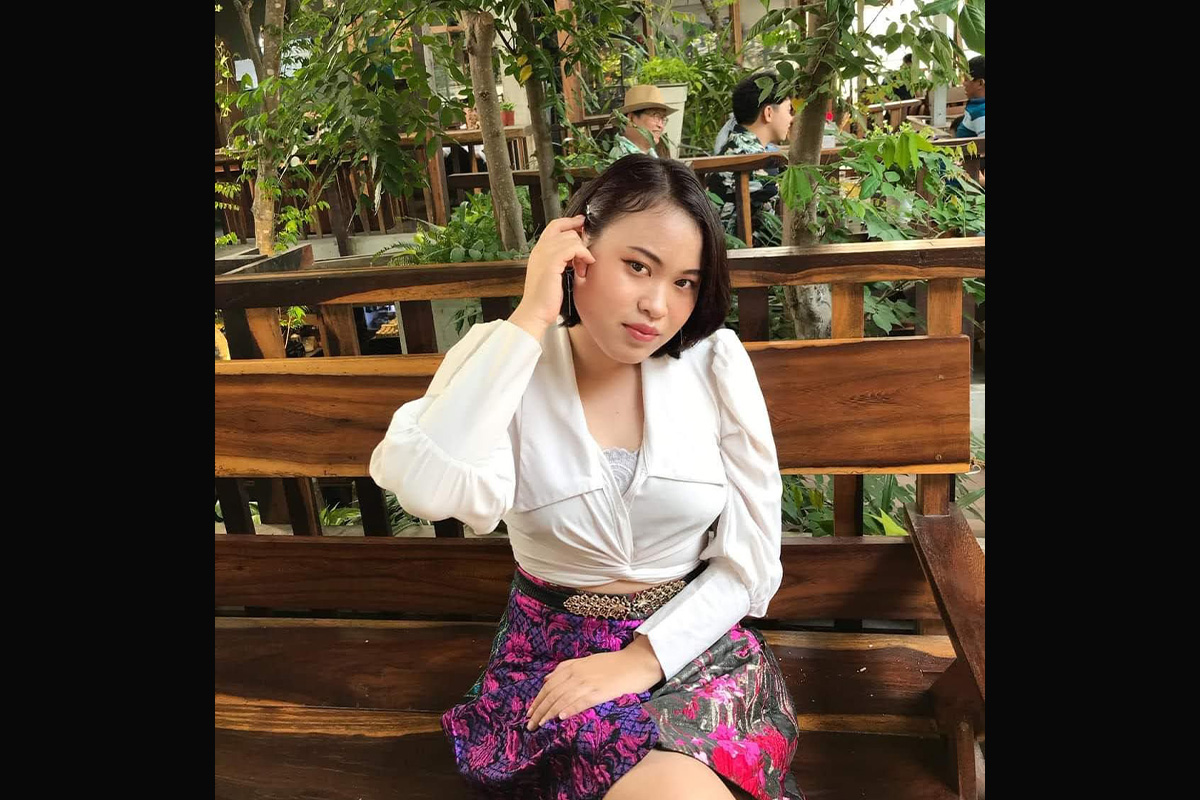 A woman sits on a wooden bench and tucks her dark hair behind her ear