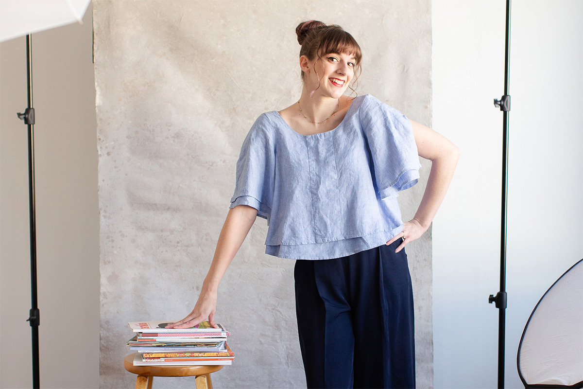 A woman stands in front of a photo backdrop, leaning onto a stack of books placed on a wooden stool