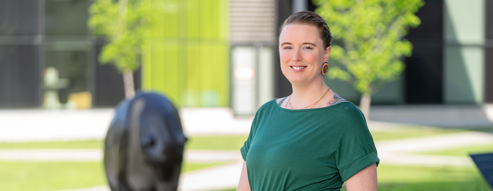 Allyson Brimstone wears a green dress and stands in front of the bear sculpture outside of Building 9, holding Cree language books