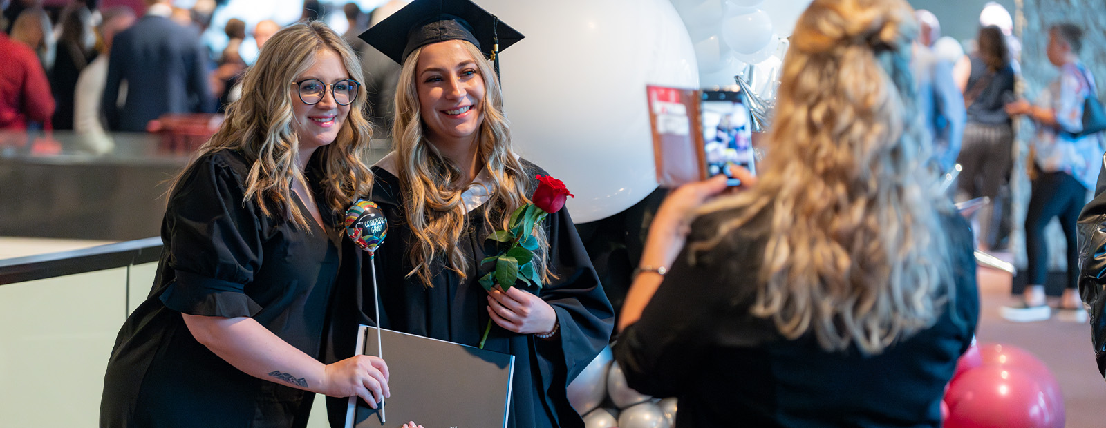 A graduate poses in her cap and gown with her mother, while a friend takes a photo of the pair with her cell phone.