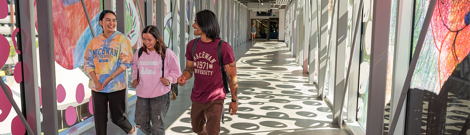 Three students wearing MacEwan-branded shirts walk across the pedway toward Allard Hall. There is an art installation on the windows of the pedway that reflects onto the floor.