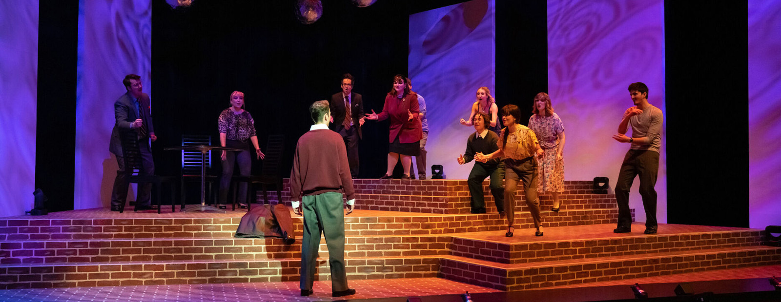 A group of actors stand on stage together with disco balls over their heads and purple lighting on the back wall
