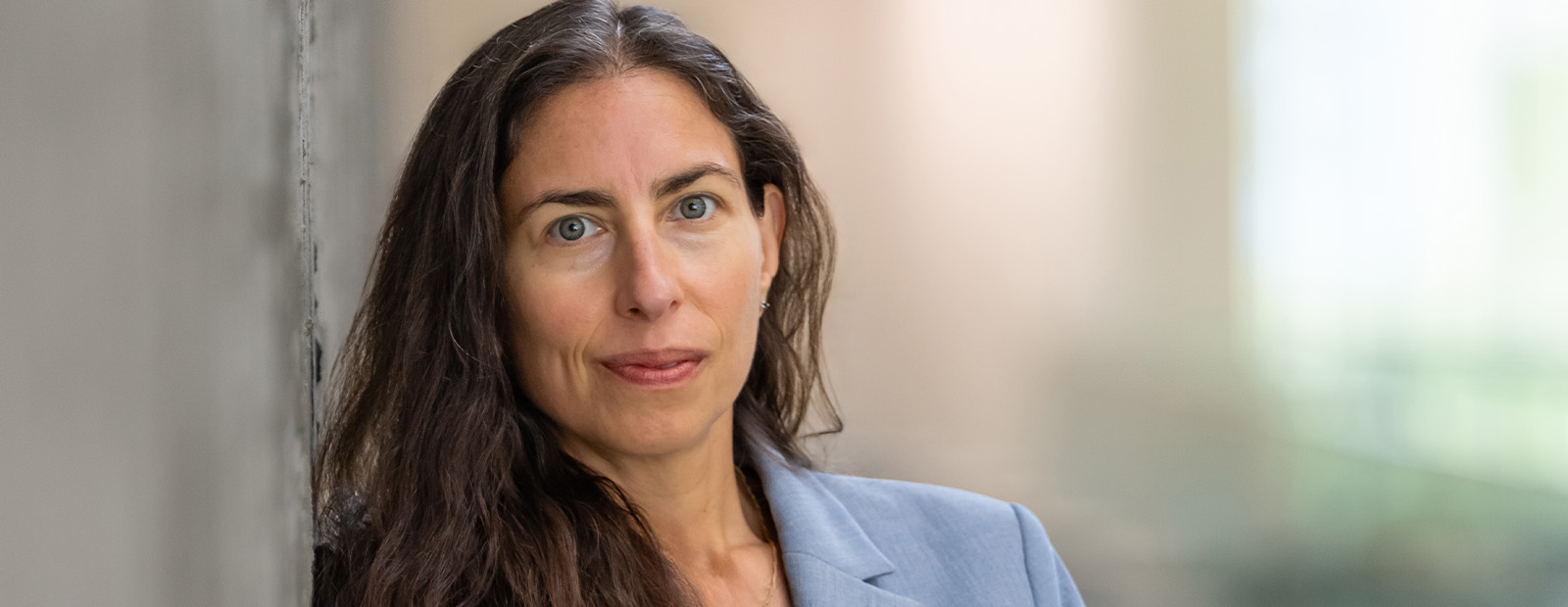 Dr. Lisa Rochman wears a light blue blazer and leans against a concrete wall in Allard Hall. She has long, brown hair and looks toward the camera with a slight smile.