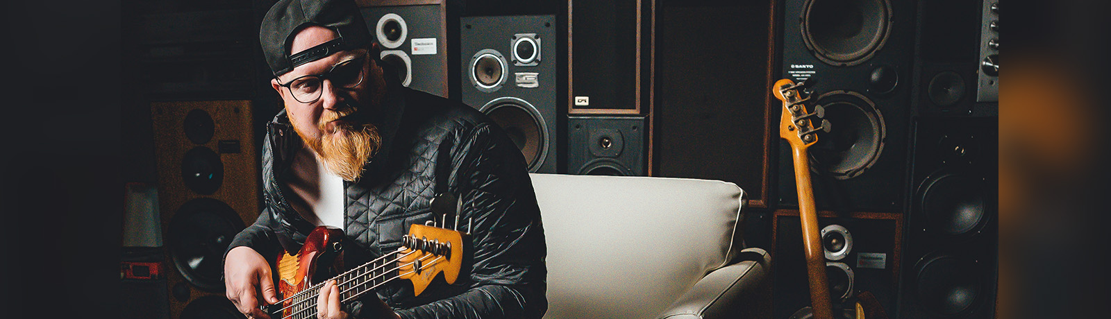 Travis Switzer sits in his studio holding his bass and wearing a black ball cap and leather jacket while holding his bass