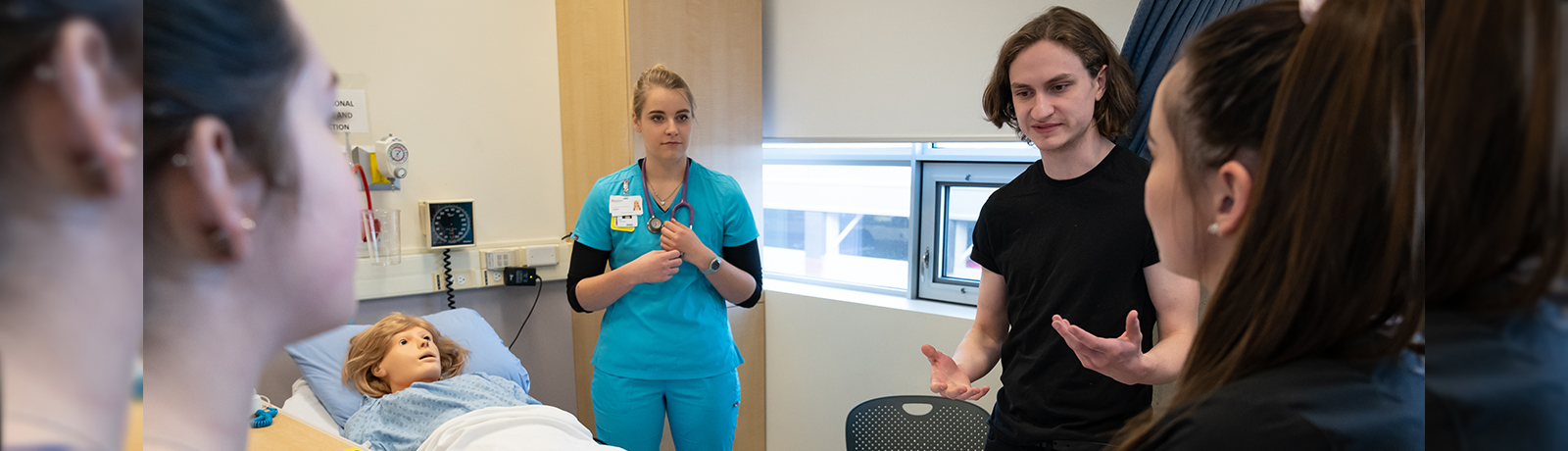 Three MacEwan students stand around a hospital bed with a nurse dressed in green scrubs. They talk while examining a training mannequin in the hospital bed.