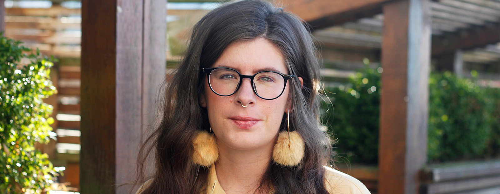 A headshot of Jessica Johns. She has dark brown hair, wears glasses, a yellow shirt and matching yellow earrings with pompoms
