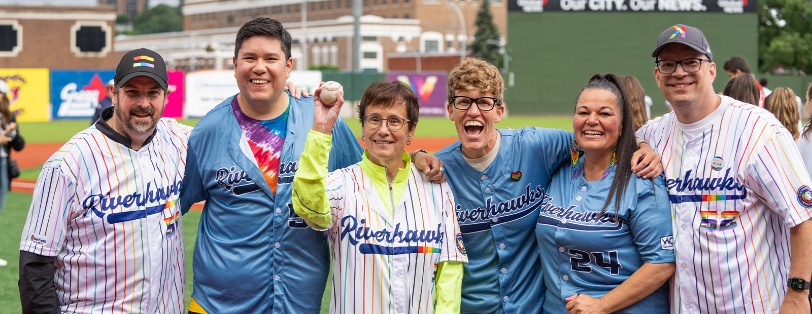 A group of people stand arm-in-arm together, with Dr. Annette Trimbee in the centre holding a baseball. All wear pride-themed Riverhawks jerseys.