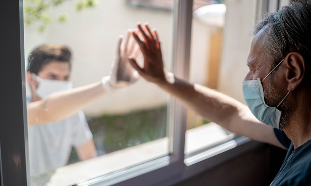 An older adult wearing a mask holds his hand up against a window while a younger person matches his hand from the other side