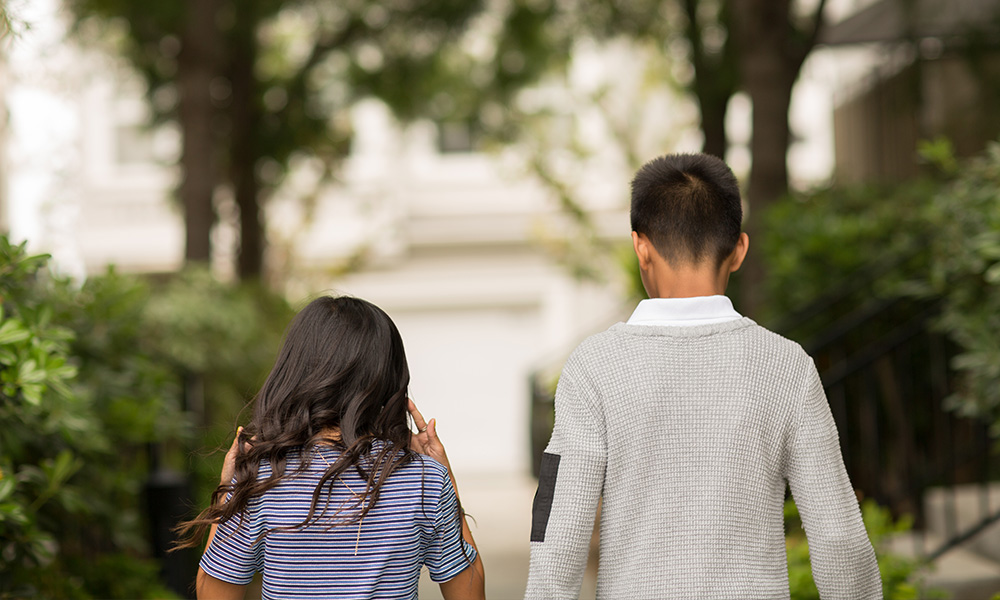 Two children walk away from the camera along a sidewalk lined with shrubs