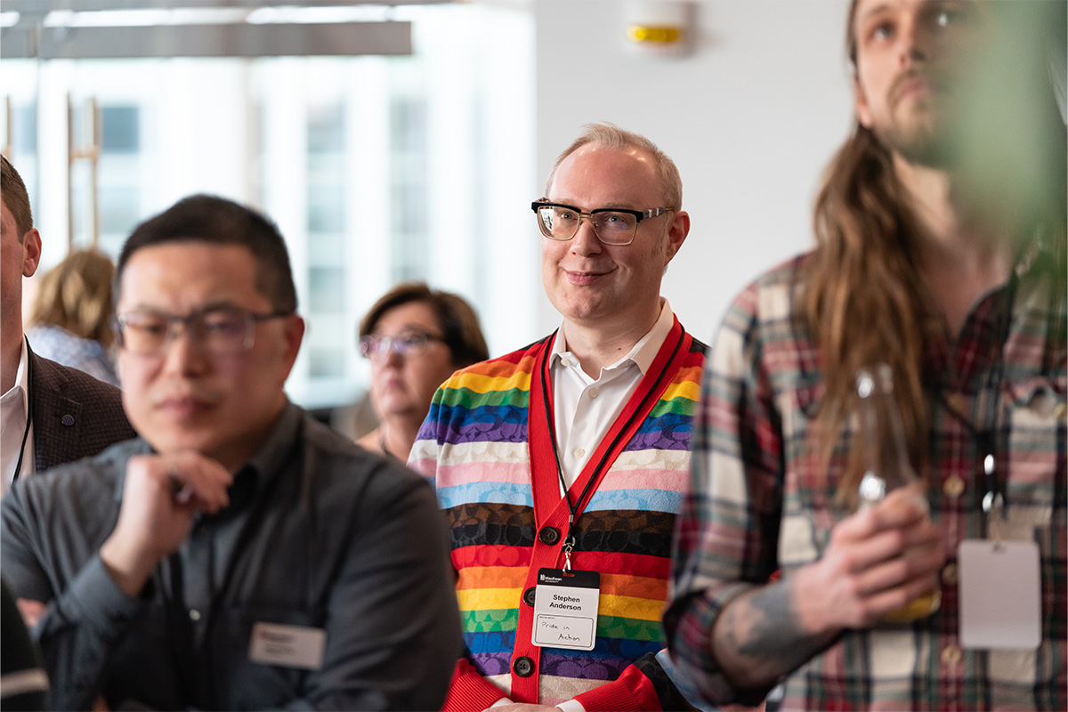 Stephen M.L. Anderson wears black-framed glasses and a rainbow-striped sweater and watches a speaker at a donor appreciation event.