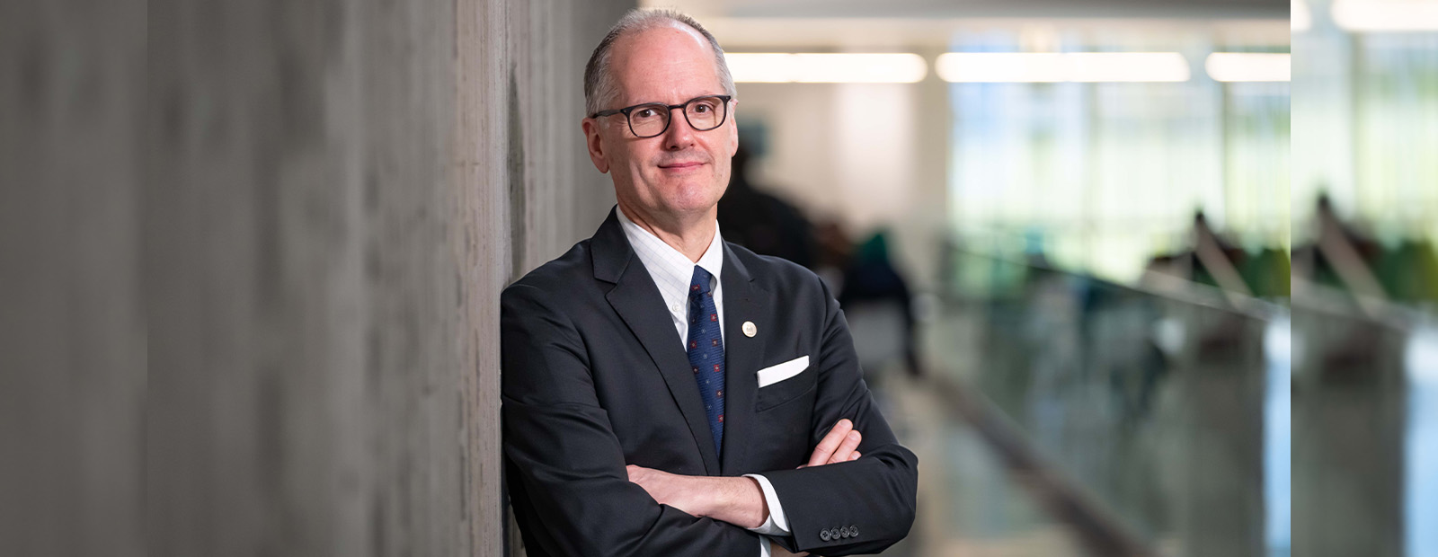 Dr. Monk wears a blazer and leans against a concrete wall in Allard Hall, his arms folded across this chest, smiling.