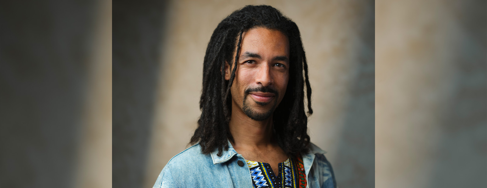 A headshot of AJA Louden, who has black dreadlocks and wears a colourful shirt beneath a denim jacket, standing in front of a grey backdrop.
