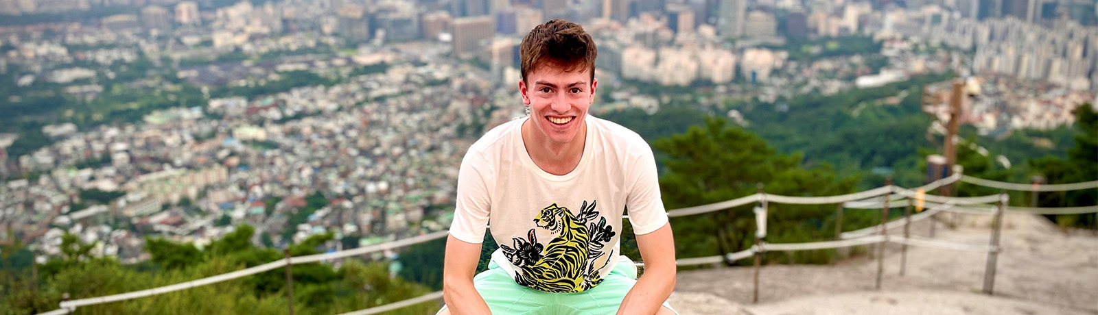 A student crouches down on a mountaintop with Seoul, Korea in the background.
