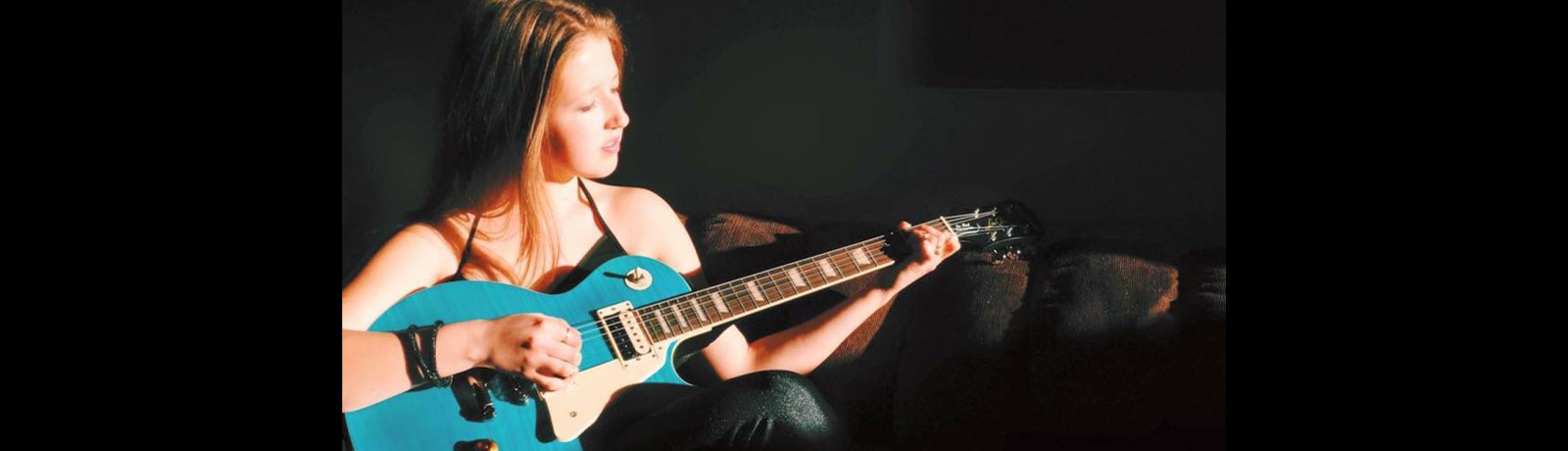 Paulina Morgan sits on a dark leather couch, playing a bright blue guitar.