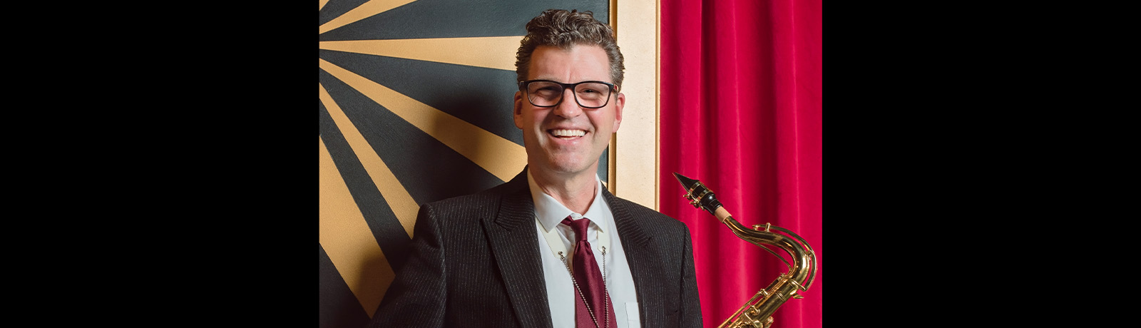 Dave Babcock holds a saxophone and stands in front of a red curtain.