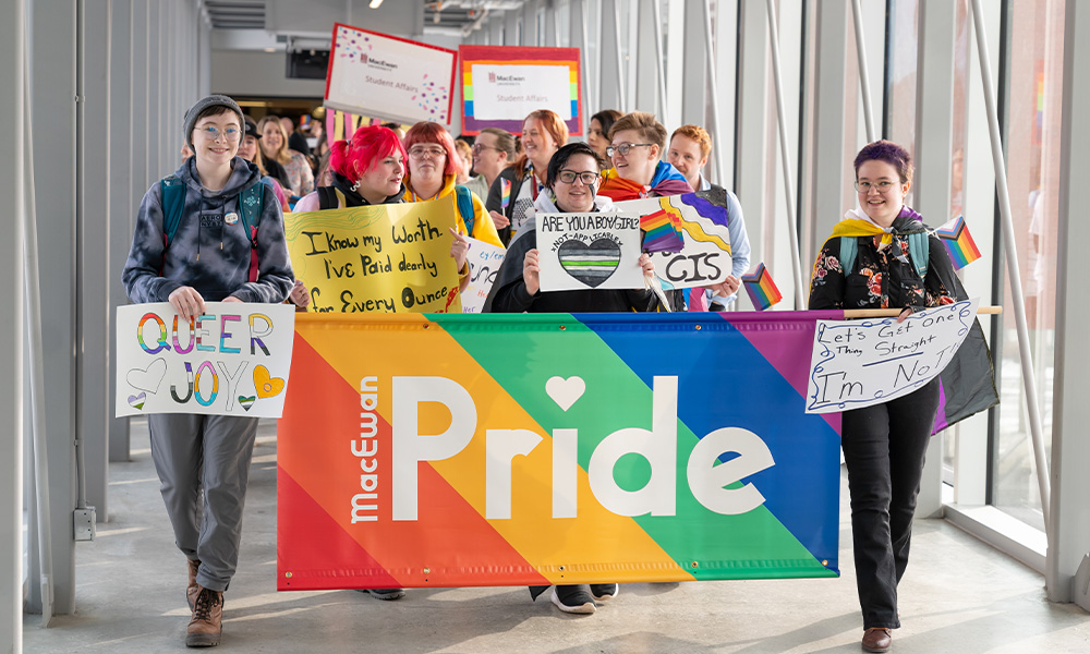 A group of students, faculty and staff walk together across the pedway into Allard Hall, holding brightly coloured signs and a rainbow Pride banner as part of MacEwan's 2023 Pride March.