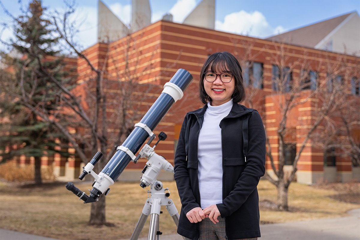 Vy Tran stands outside of Building 5 on campus next to a telescope.