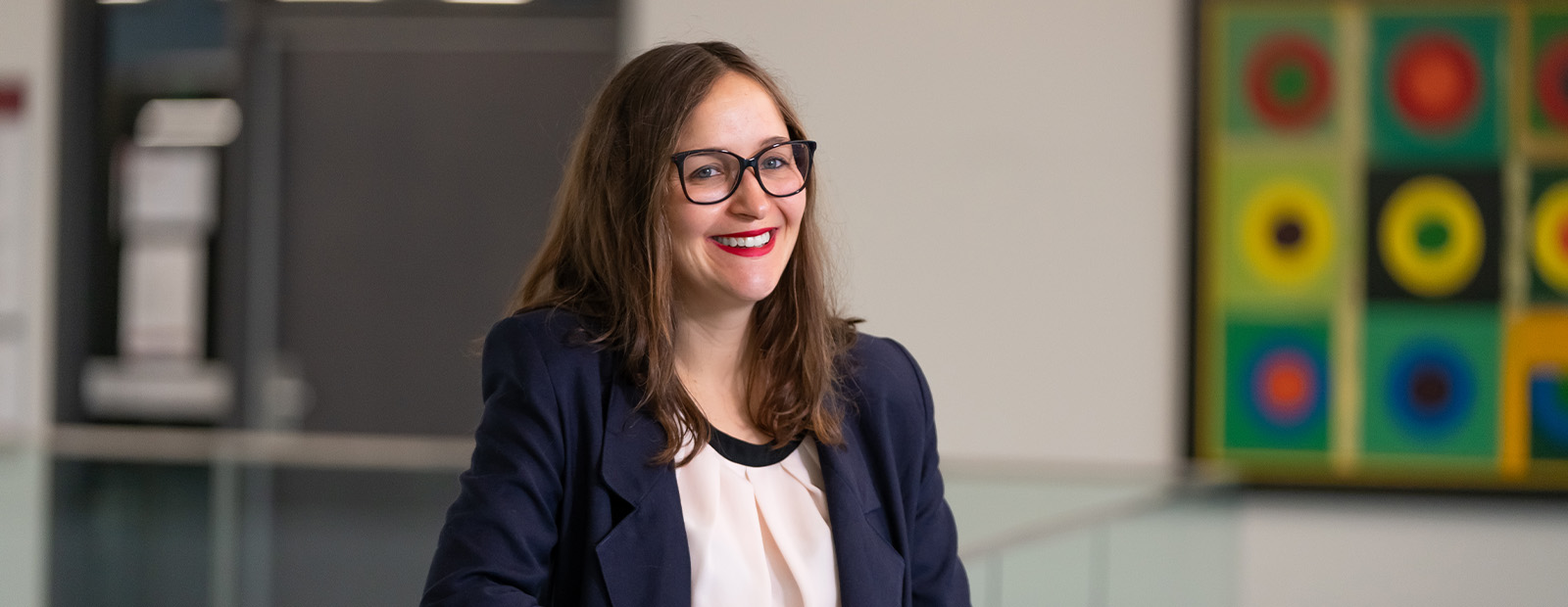 Dr. Papin wears a blue blazer over a white shirt and leans against a glass railing in Allard Hall, smiling.