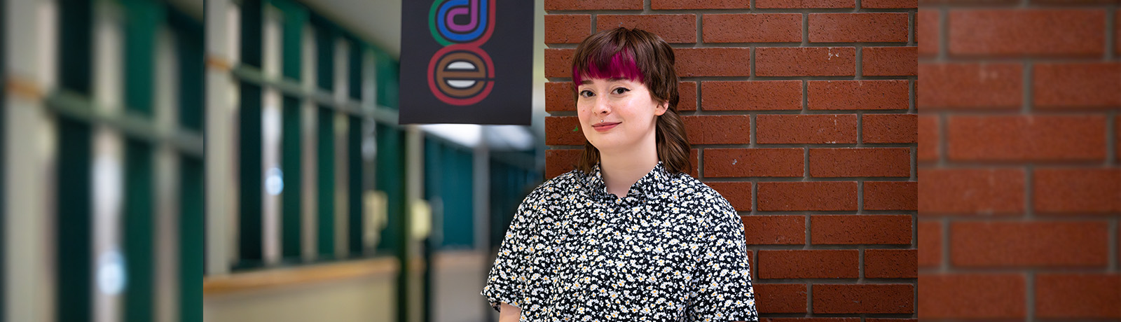 Alana Whitson has brown and magenta hair and wears a black and white floral shirt, standing in front of a Pride banner in Building 7.