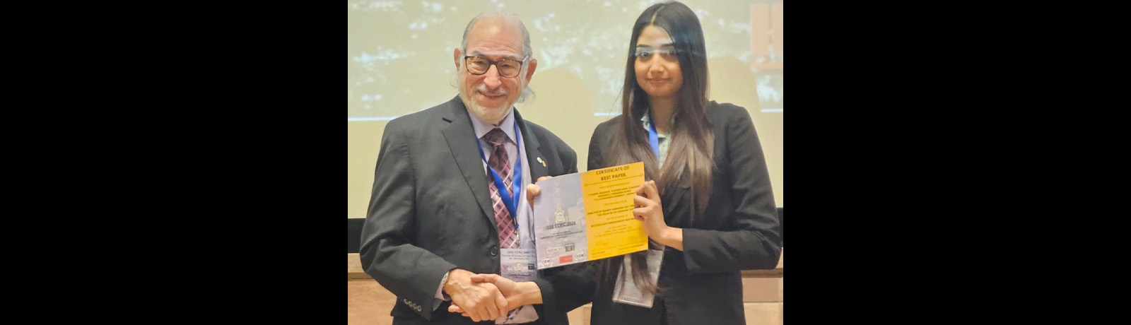Ayesha Khan receives the Best Paper award on stage, presented by a man in a black suit.