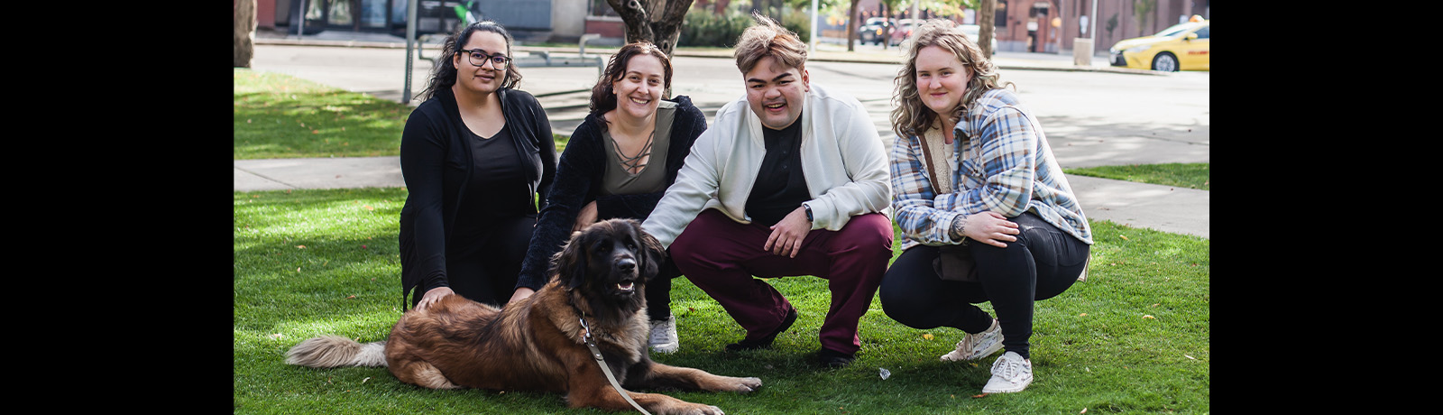 Four people crouch on the grass with a large brown and black dog, all smiling.