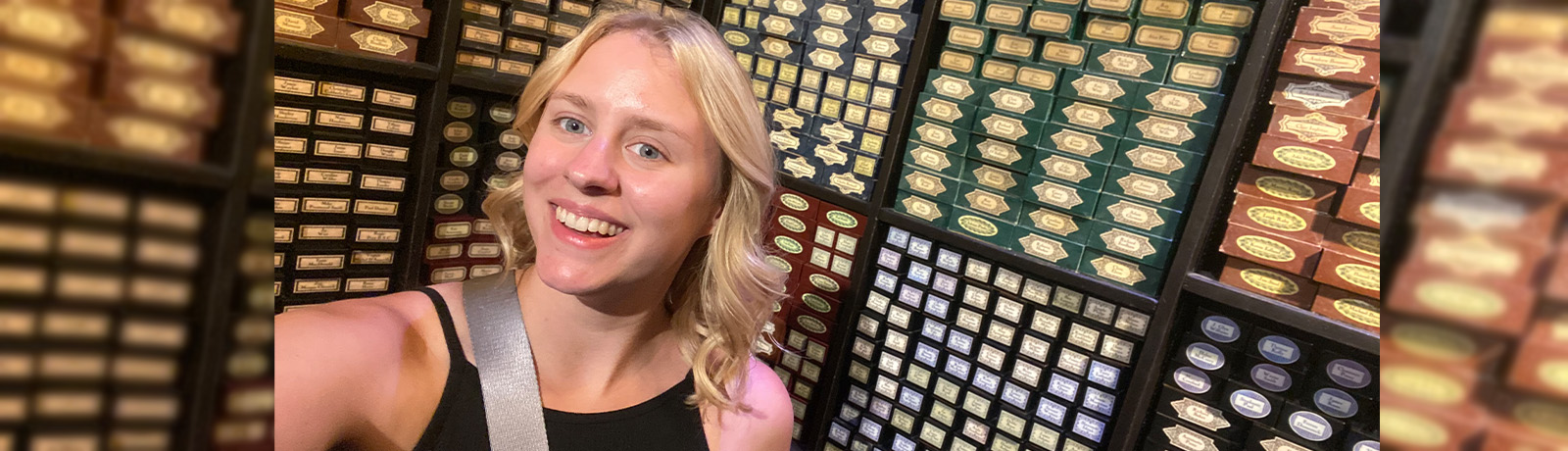 Brynne, a young woman with blonde hair and blue eyes, smiles at the camera in front of a wall displaying many small boxes of items.