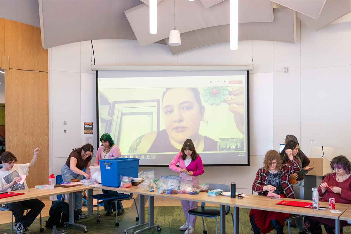 Brittany Johnson demonstrates beadwork on a large screen in a classroom while gathered MacEwan community members watch and follow along.