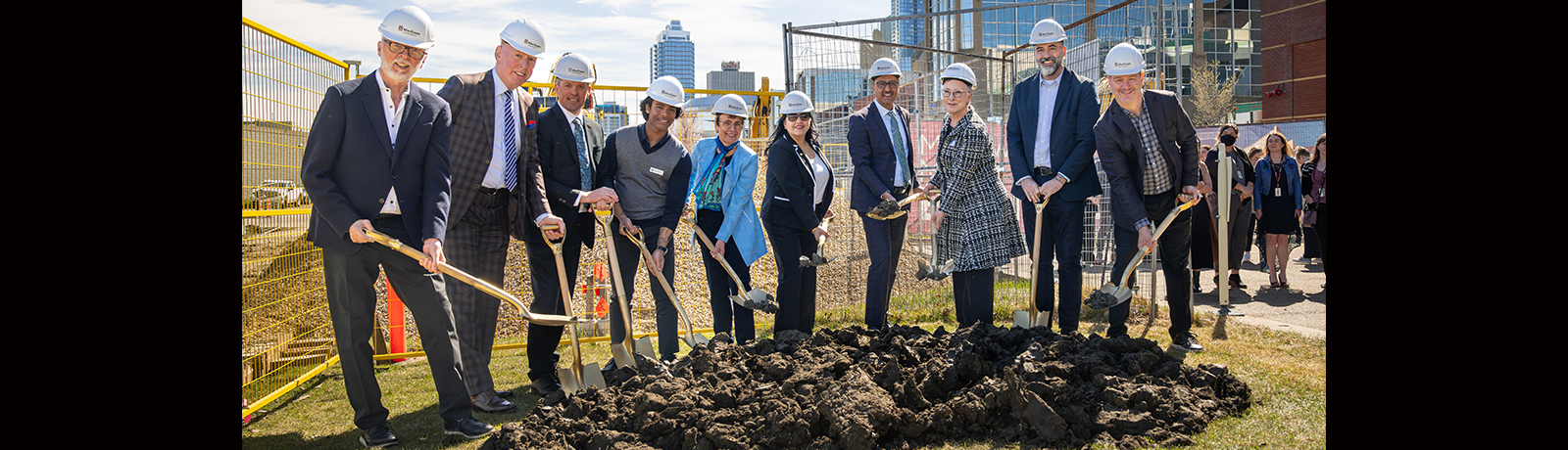People standing in a semi-circle around a pile of dirt, each holding a shovel and wearing a hard hat.