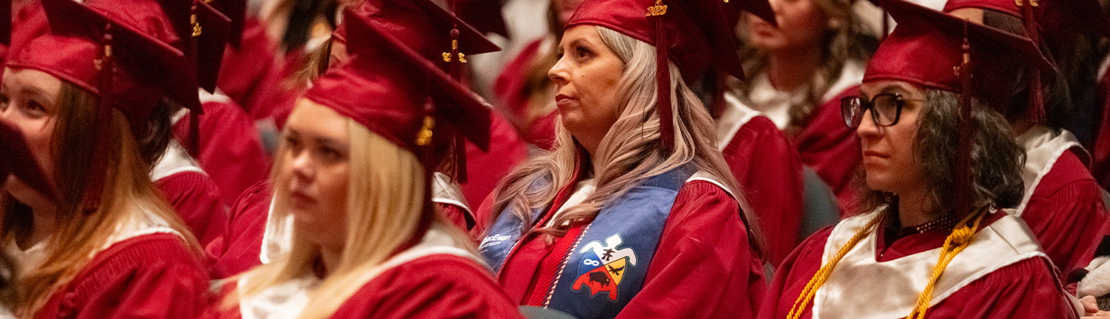 Graduates sit in red caps and gowns, watching the convocation ceremony. One wears an Indigenous stole and another wears a yellow cord.