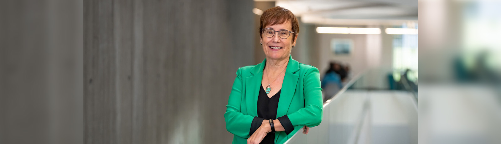Dr. Annette Trimbee wears a green blazer and leans against a glass railing in Allard Hall.