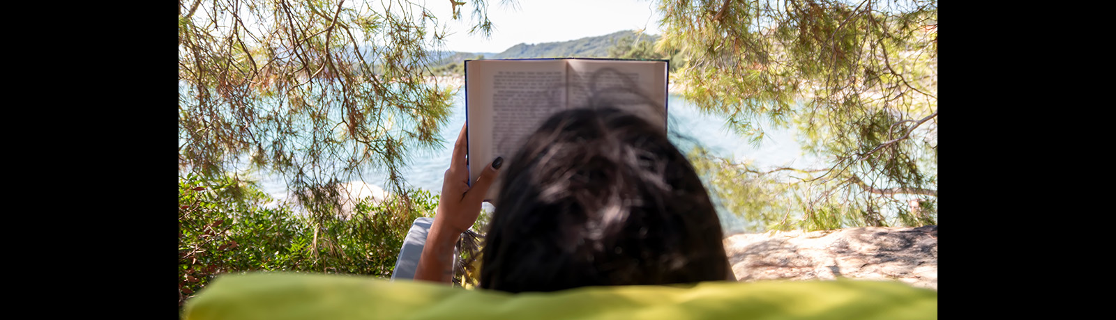 A woman reads a book and faces out toward a body of water
