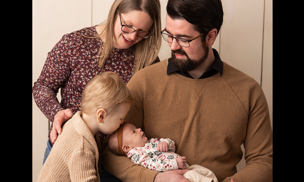 A man in a brown sweater and glasses holds a newborn baby while a blonde woman looks over his shoulder at the baby.