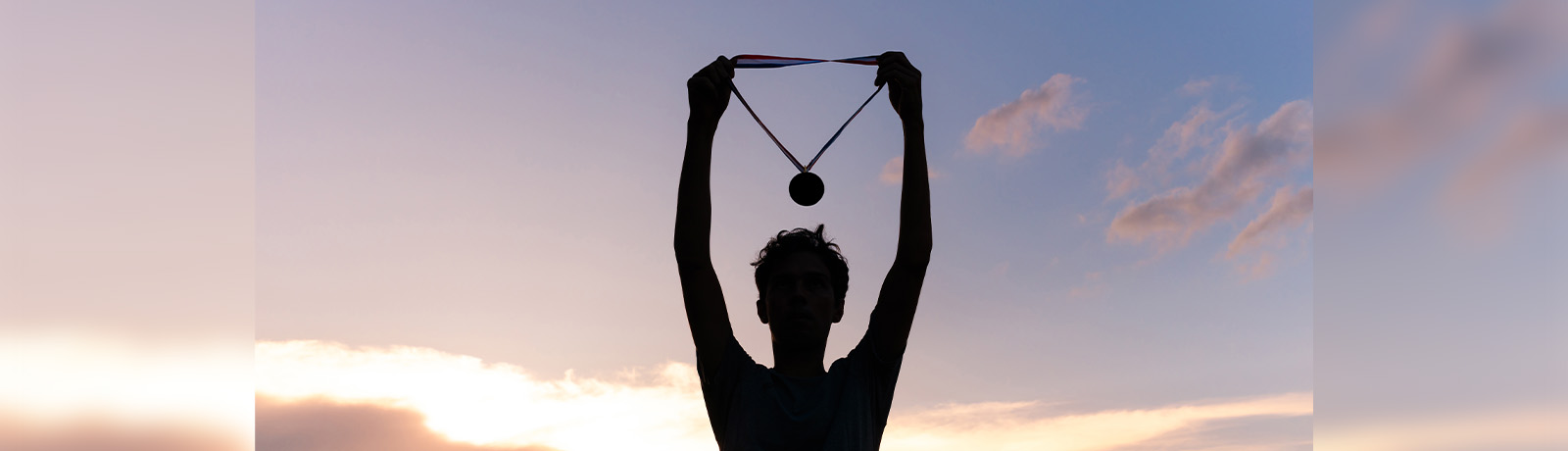 An athlete stands in shadow holding up a medal in front of a sunset.