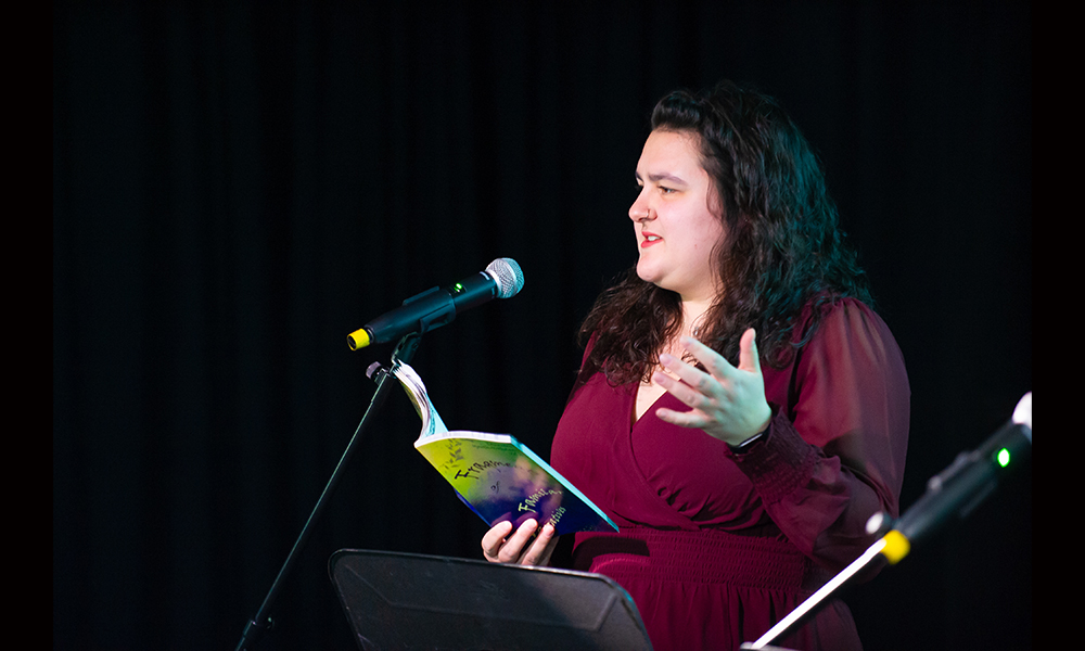 Kyra Droog is standing at a microphone reading from a book