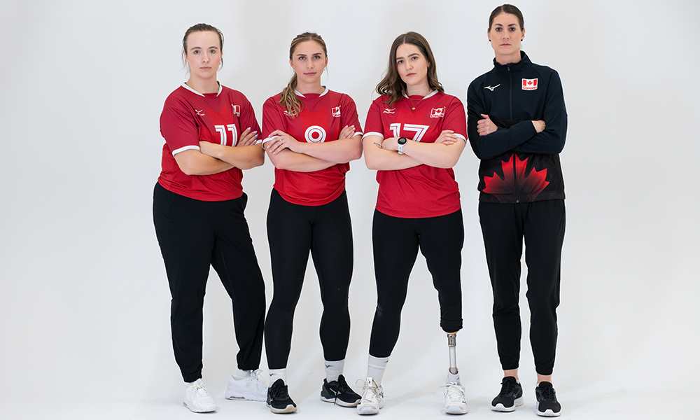 Members of the Paralympic Sitting Volleyball team stand in front of a white background with their arms crossed. Three players wear their team uniforms of red shirt and black pants. The coach wears an all-black uniform. 