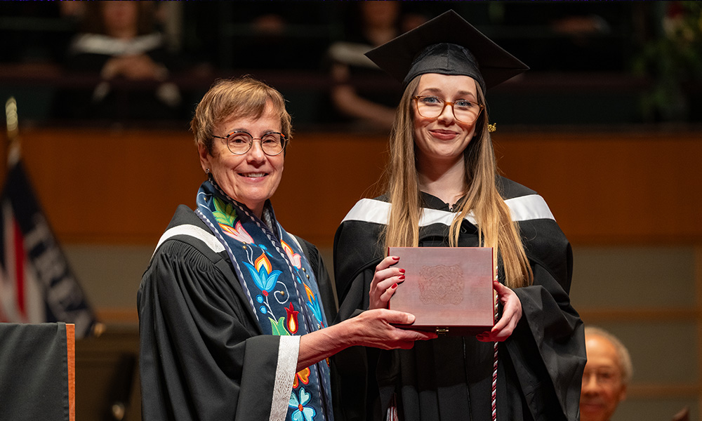 Dr. Trimbee and Eva Hollas hold a wooden box containing the president's medal, both are smiling and wearing convocation regalia