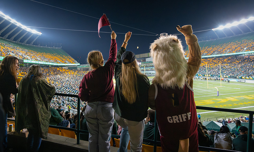 Two students cheer with the Griff mascot as they look onto the Edmonton Elks football field.