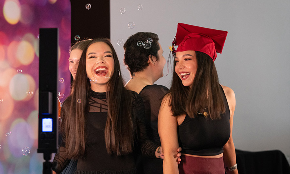 Two students, one wearing a grad cap, laugh together as bubbles float past them.
