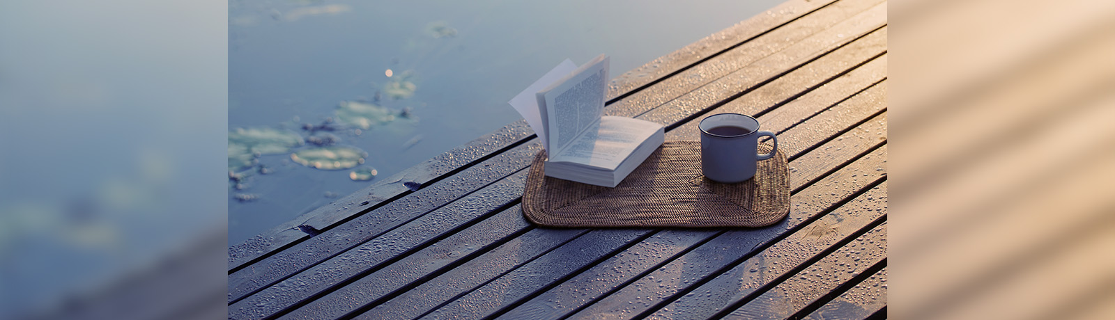 An open book and a mug of coffee on a woven placement sit on top of a dock covered in dew.