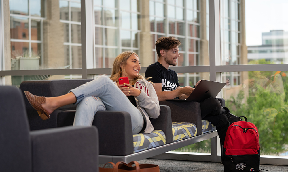 Two students lounge on seating by a large window, one looks at her phone and one is holding a laptop