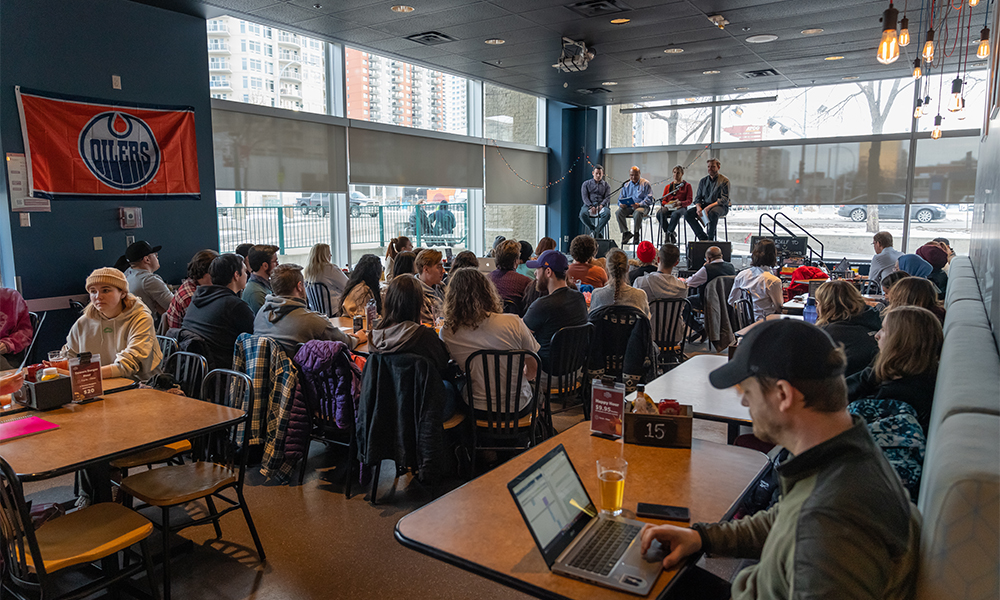 People sit at tables and some face the stage where a group of four people sit on high stools at microphones