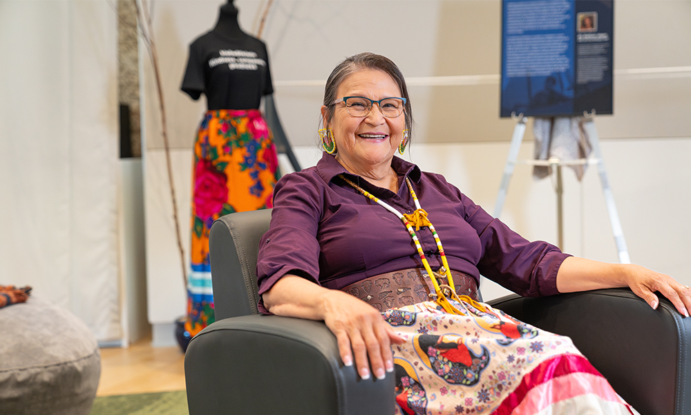 Daphne Alexis sitting in a black leather chair smiling, wearing a ribbon skirt