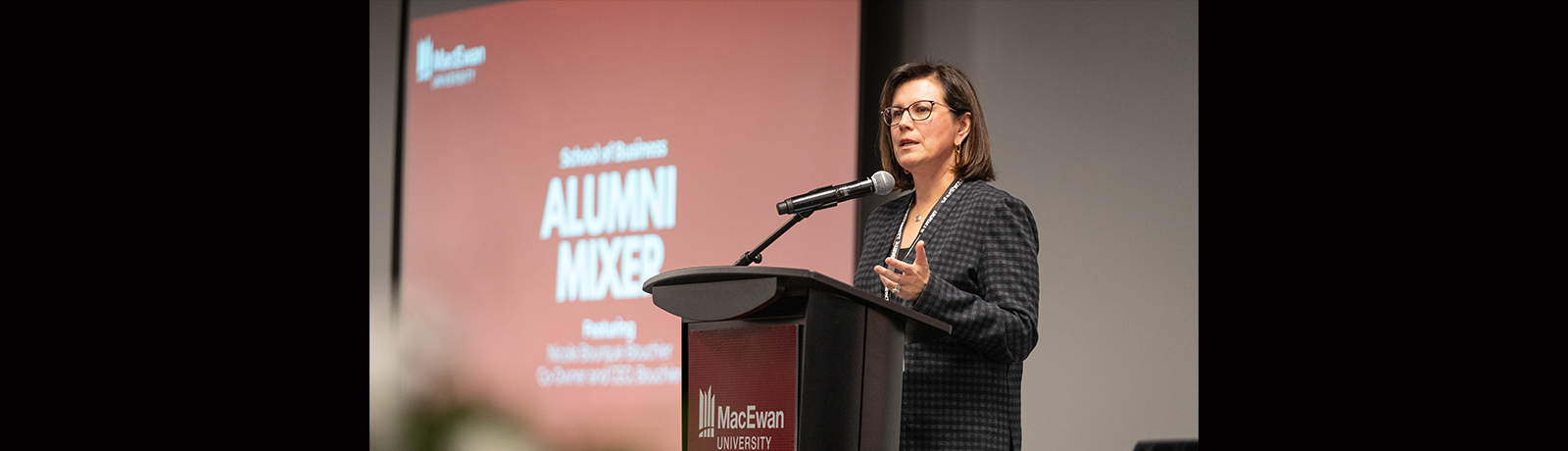 A woman stands on stage at a podium speaking, a screen behind her reads Alumni Mixer