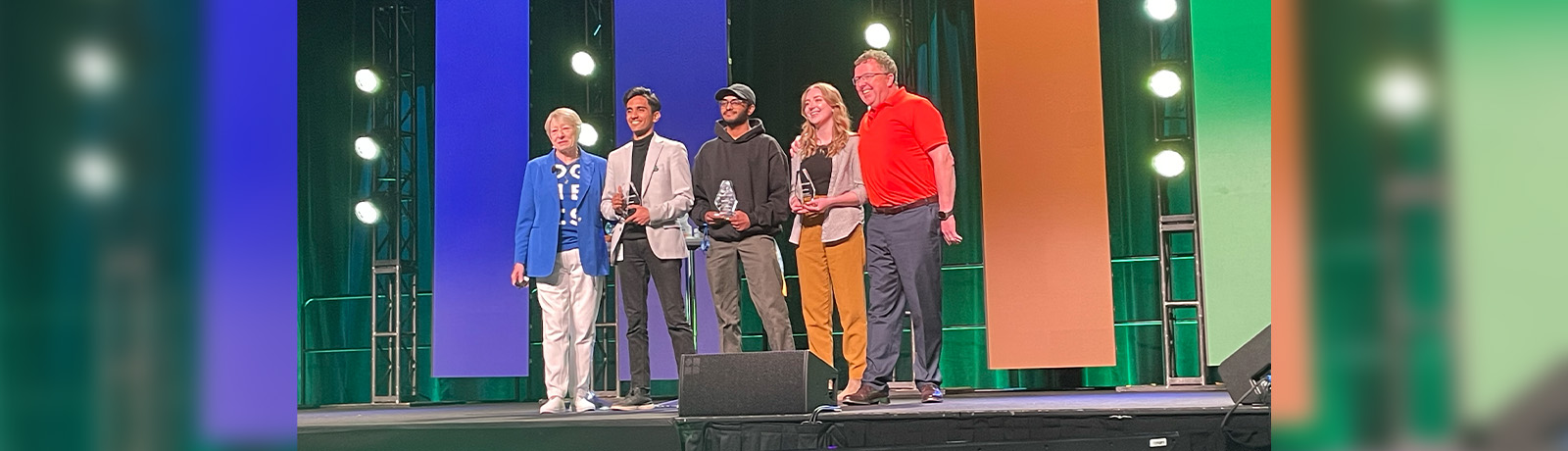 Five people stand on a stage holding awards. Sujit Velmurugan stands in the middle wearing a black hoodie.